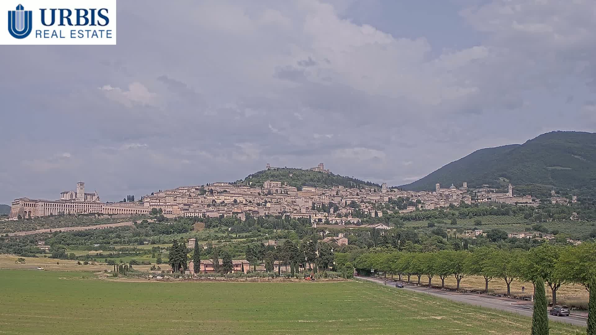 A hilltop town with many buildings and a large basilica, nestled between green hills under a partly cloudy sky.