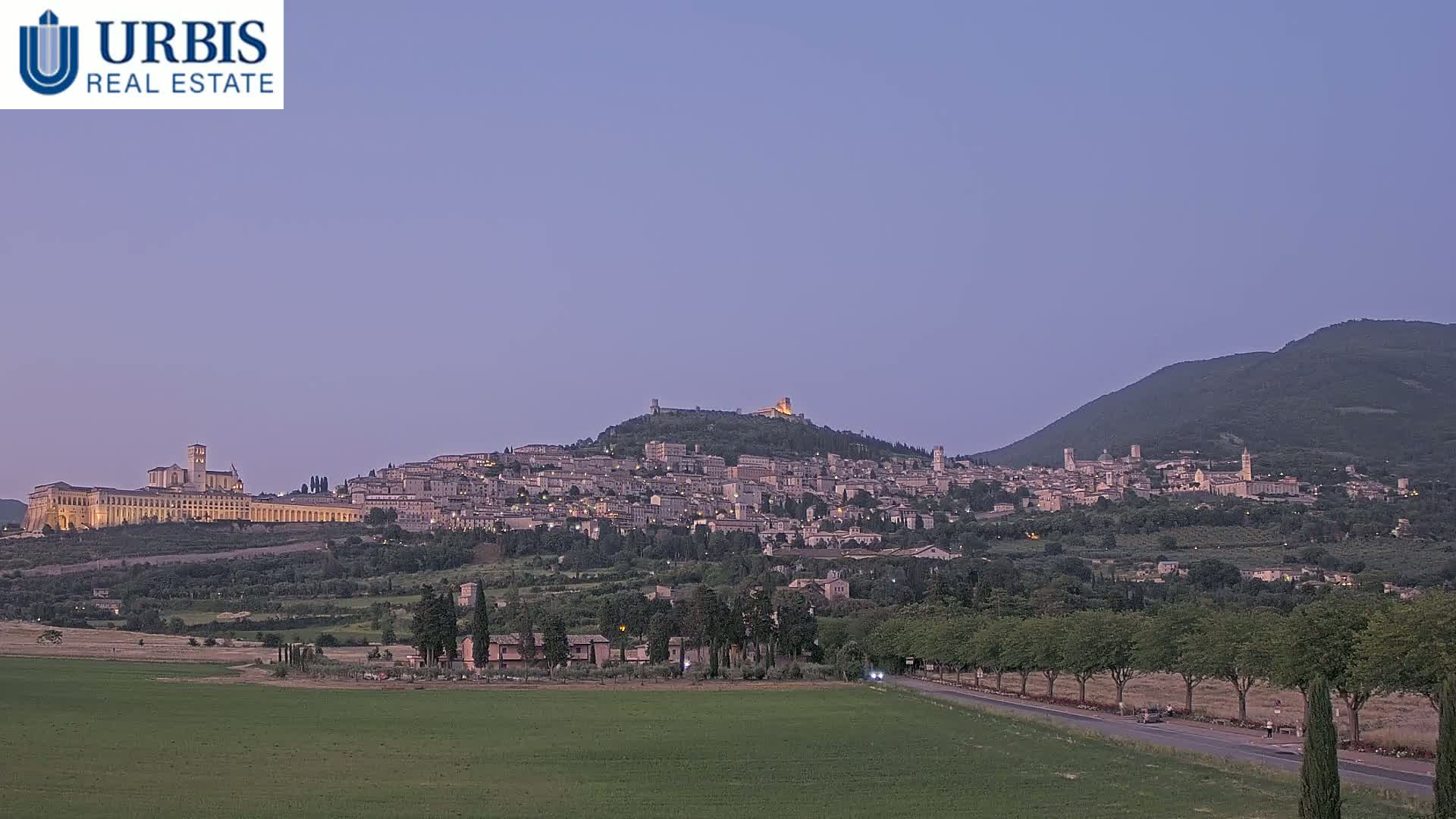 A hilltop town with a large basilica is illuminated at dusk under a pale purple sky, with a road and green fields in the foreground.