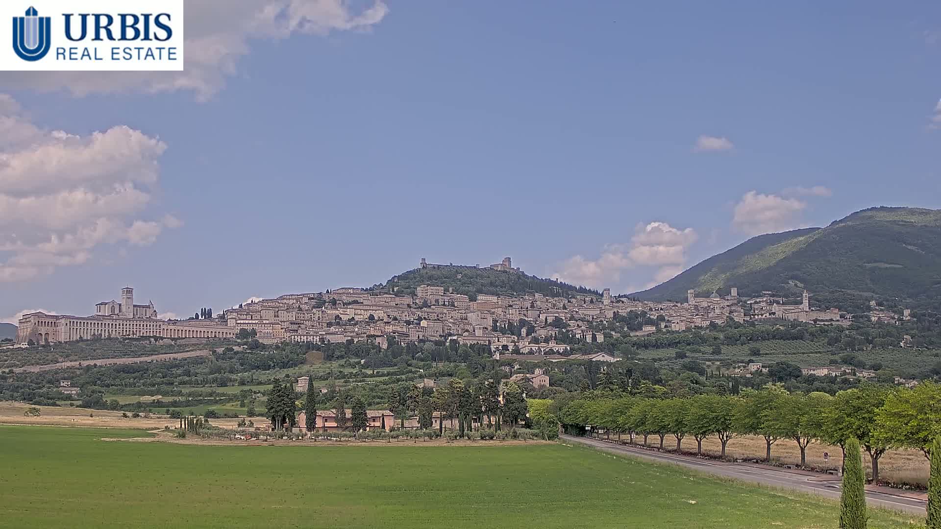 A hilltop town, nestled amongst green hills under a mostly clear sky with scattered clouds, is visible from a grassy field.