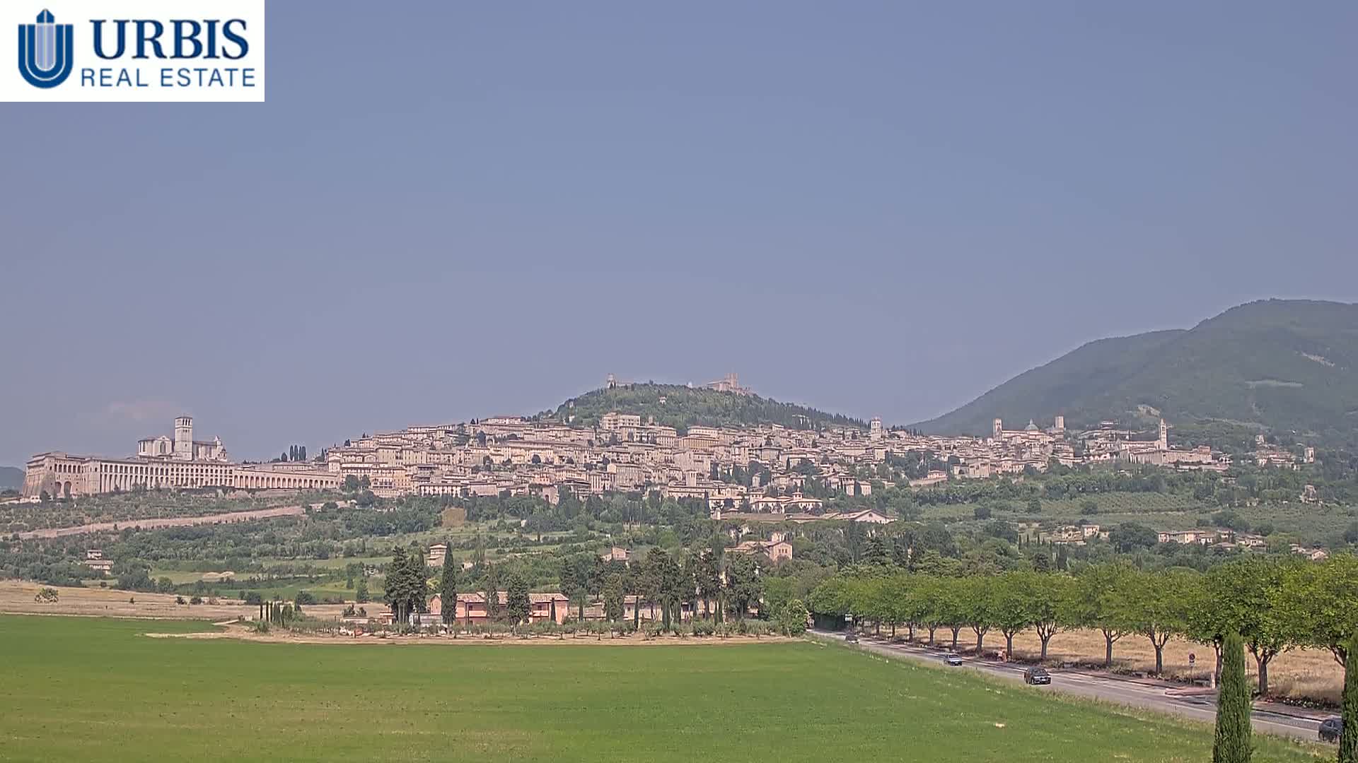 A large town is nestled on a hill under a clear blue sky, with green fields and trees in the foreground.