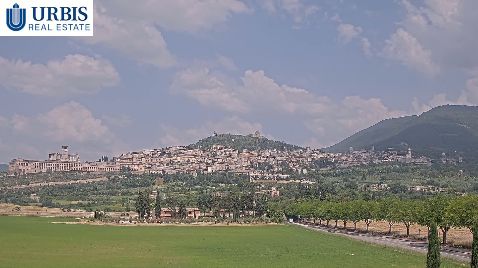 A hilltop town with numerous buildings, including a large basilica, is seen under a partly cloudy sky, with a flat green field in the foreground.