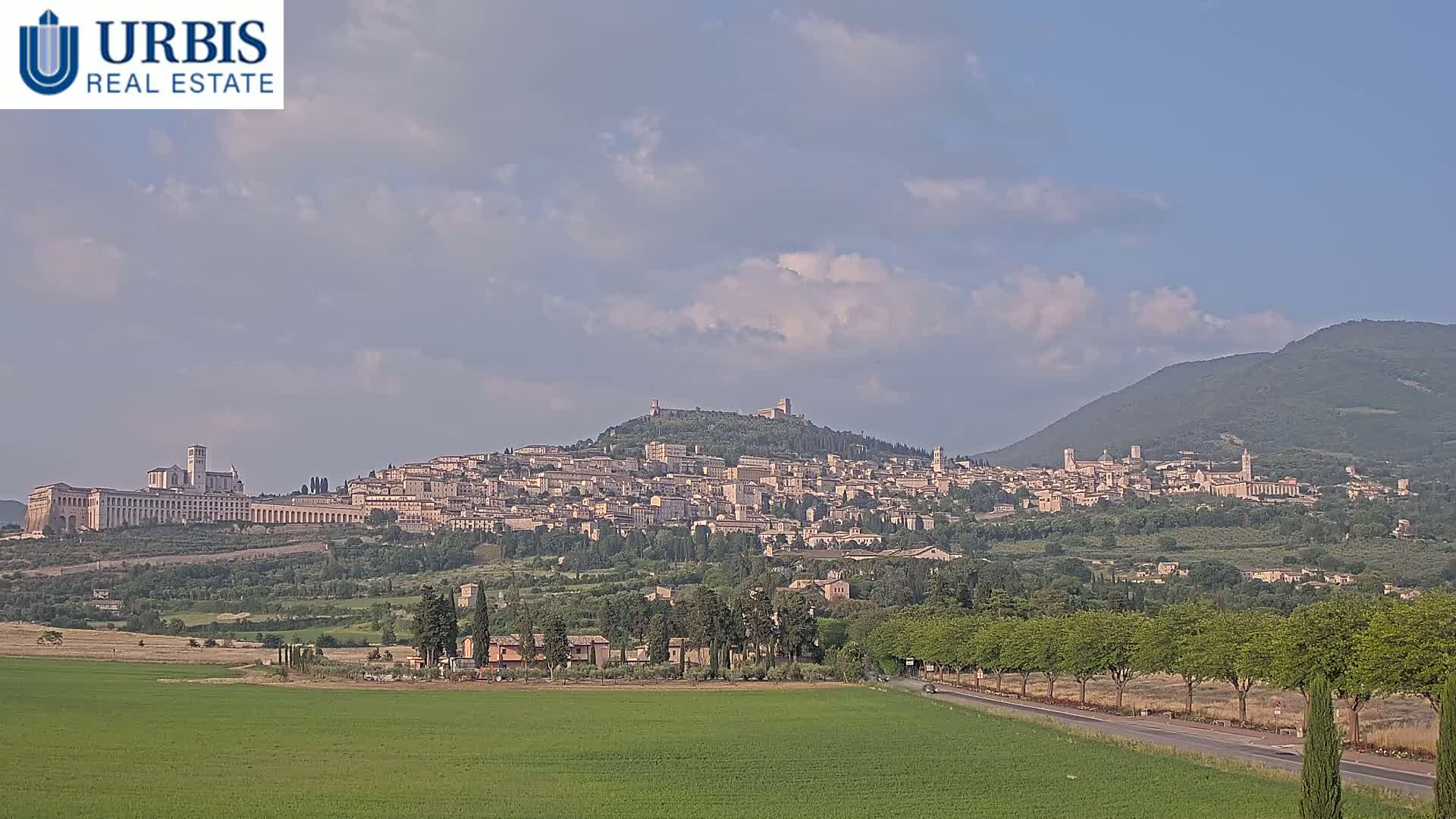 A hilltop town with numerous buildings, including a large basilica, is visible under a partly cloudy sky, with a green valley and mountains in the background.