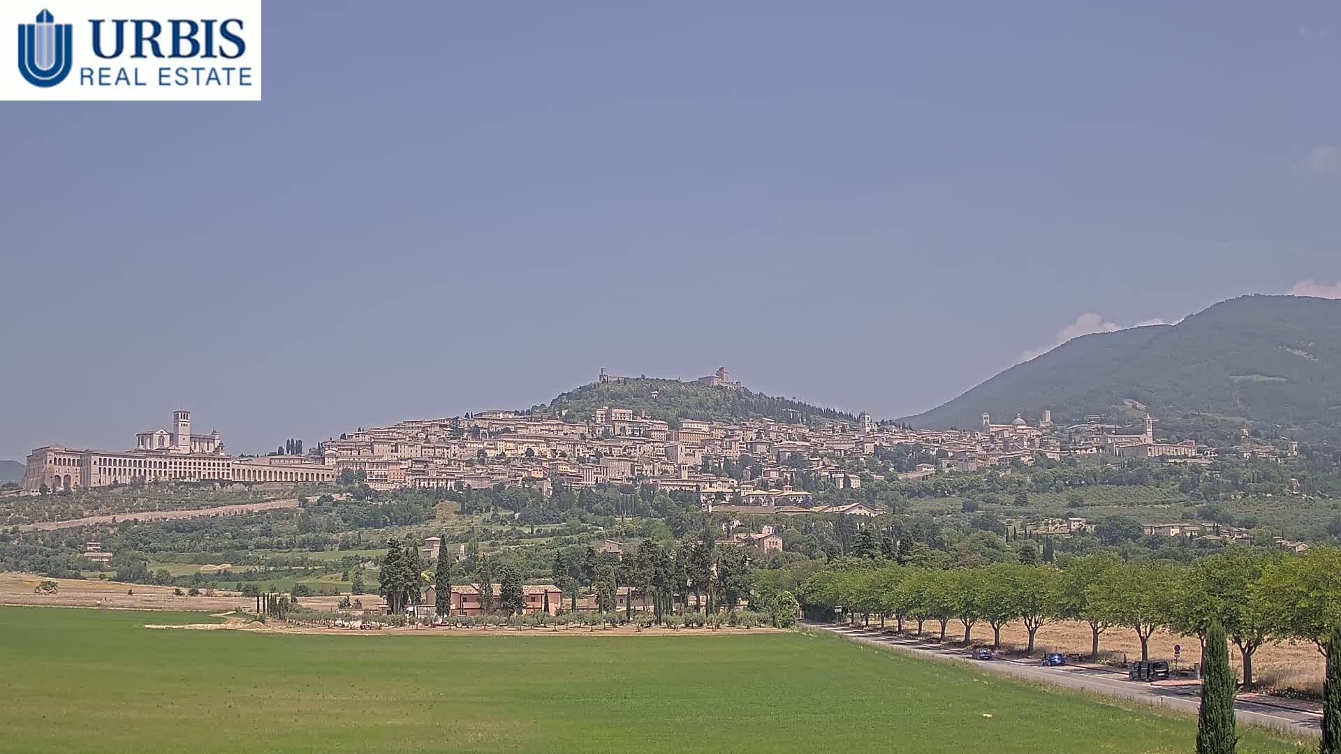 A hilltop town with many buildings, including a large basilica, is visible in the distance under a clear, bright sky, with green fields and trees in the foreground.