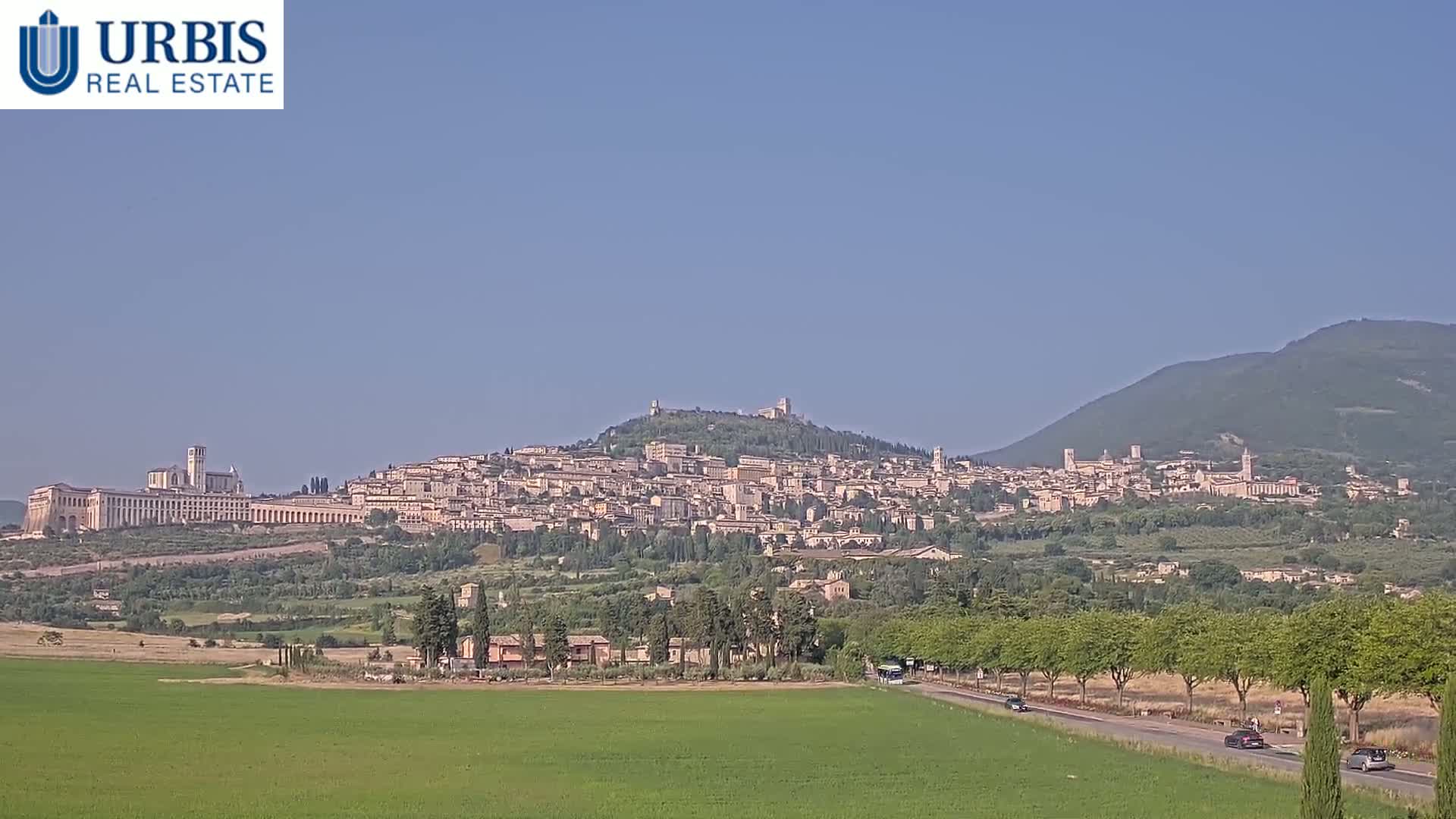 A hilltop town, possibly Assisi, is visible in the distance under a clear, sunny sky, with green fields and trees in the foreground.