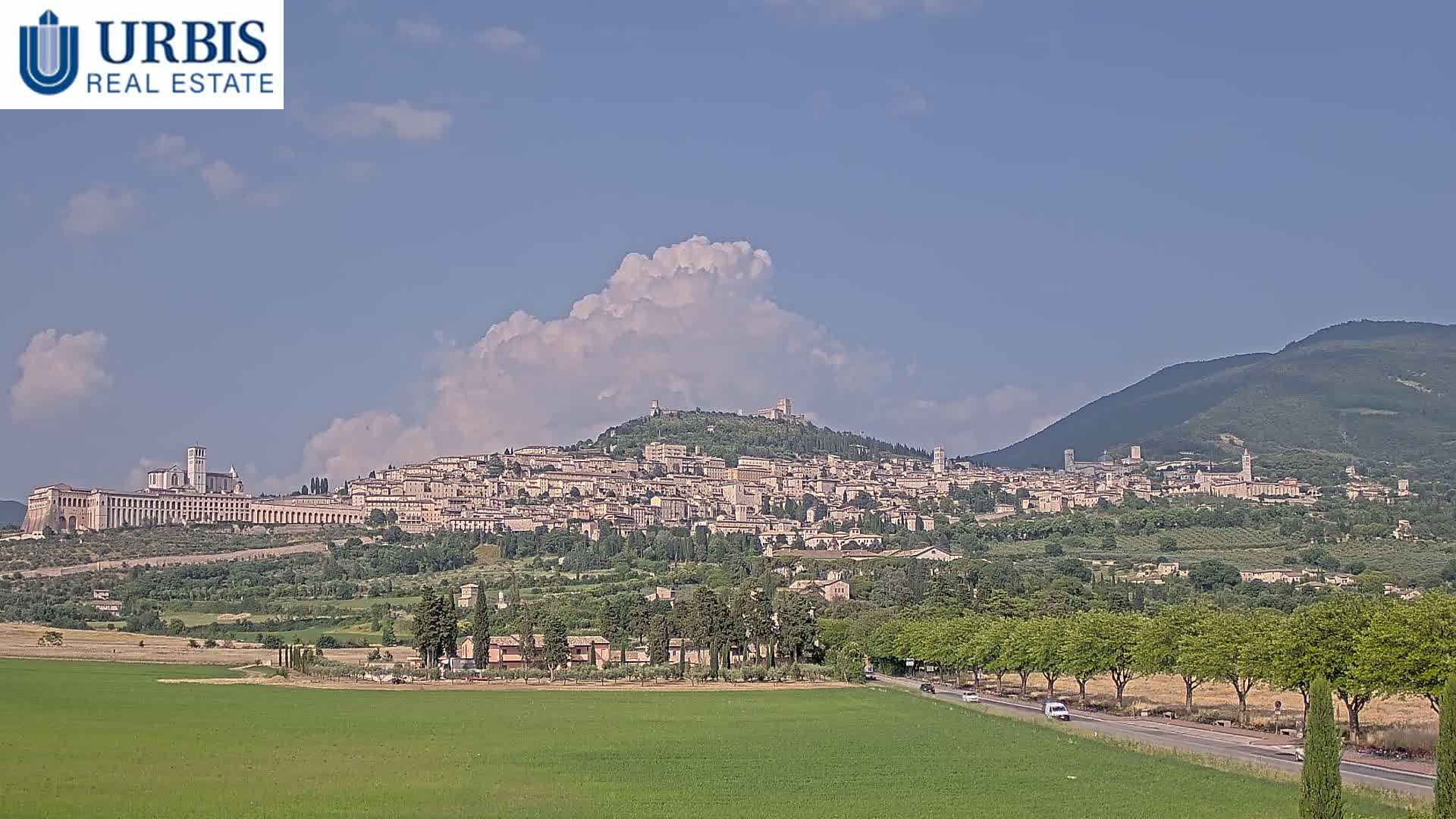 A large hill town, situated against a backdrop of mountains under a partly cloudy sky, is viewed from a distance across a green field and a road lined with trees.