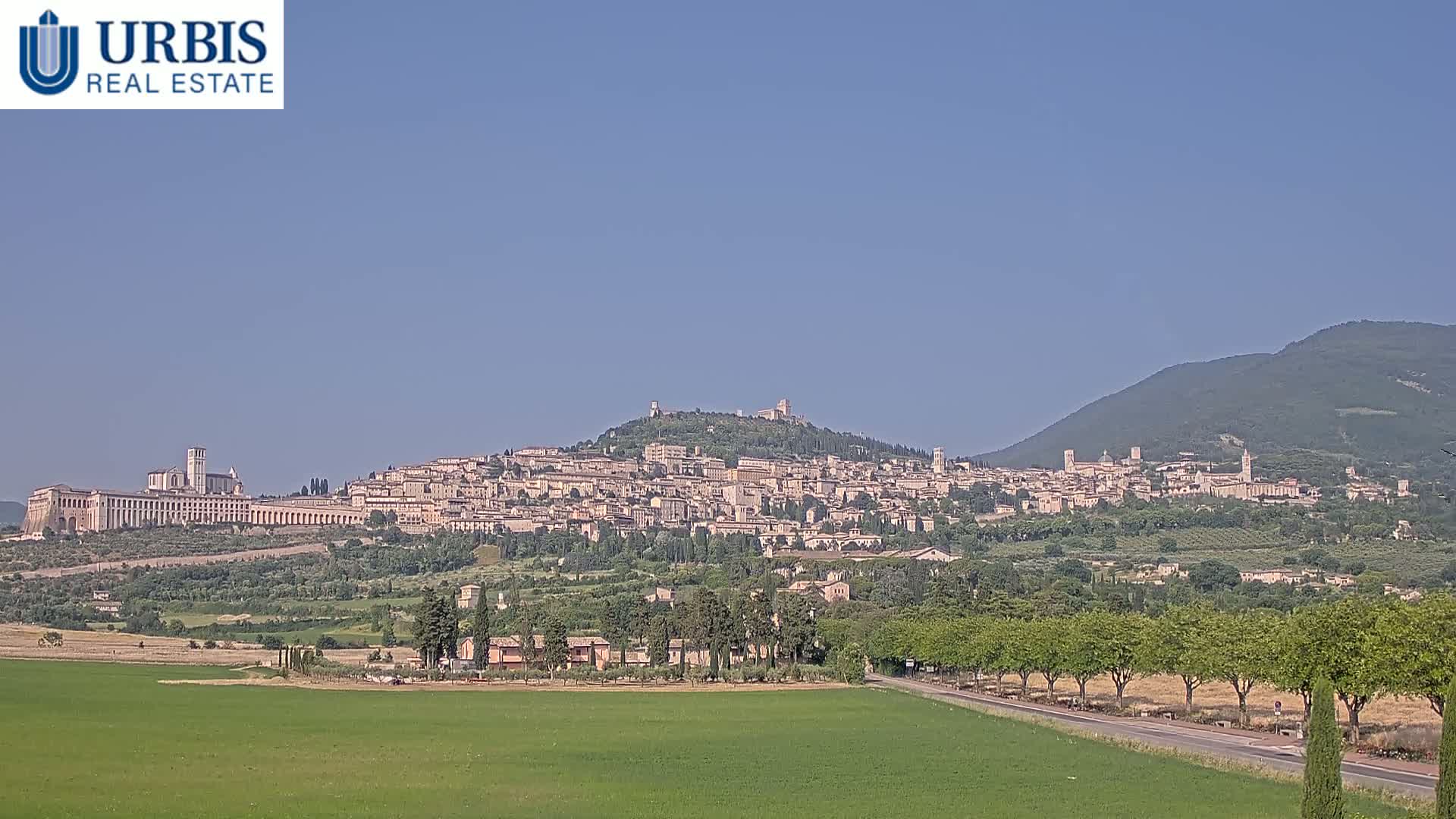A hilltop town with numerous buildings, a large basilica, and surrounding greenery is visible under a clear blue sky.