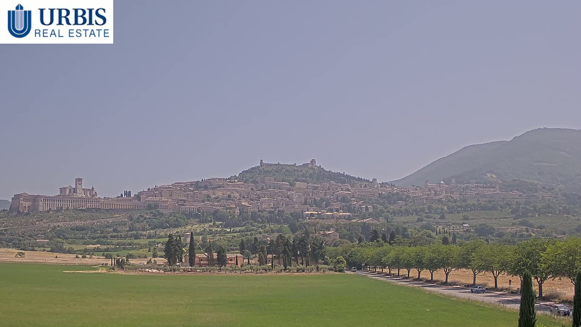 A hazy, sunny day reveals a sprawling hilltop town, with a large basilica on the left and a mountain in the background.