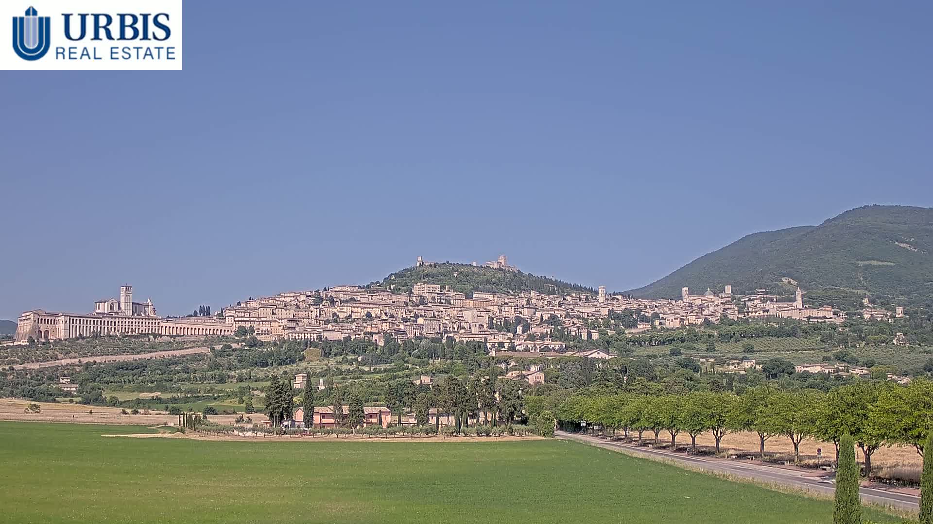 A hilltop town with numerous buildings, including a large church, is visible under a clear blue sky, with green fields and trees in the foreground.