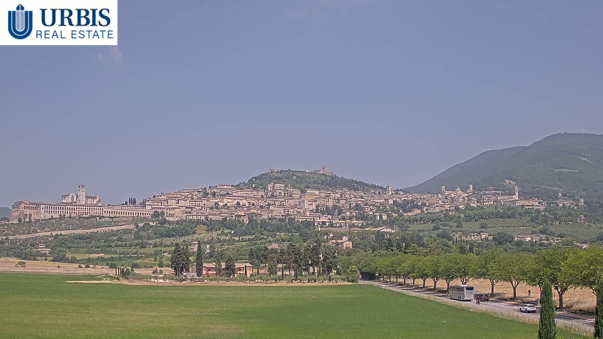 A hilltop town with numerous buildings, including a large basilica, stretches across a verdant landscape under a clear, sunny sky.