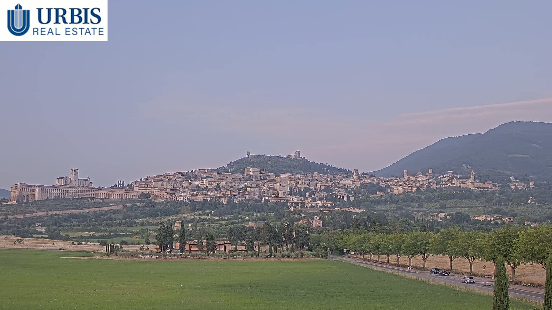 A large hilltop town, nestled between rolling green hills under a pale, clear sky, is viewed from a distance.