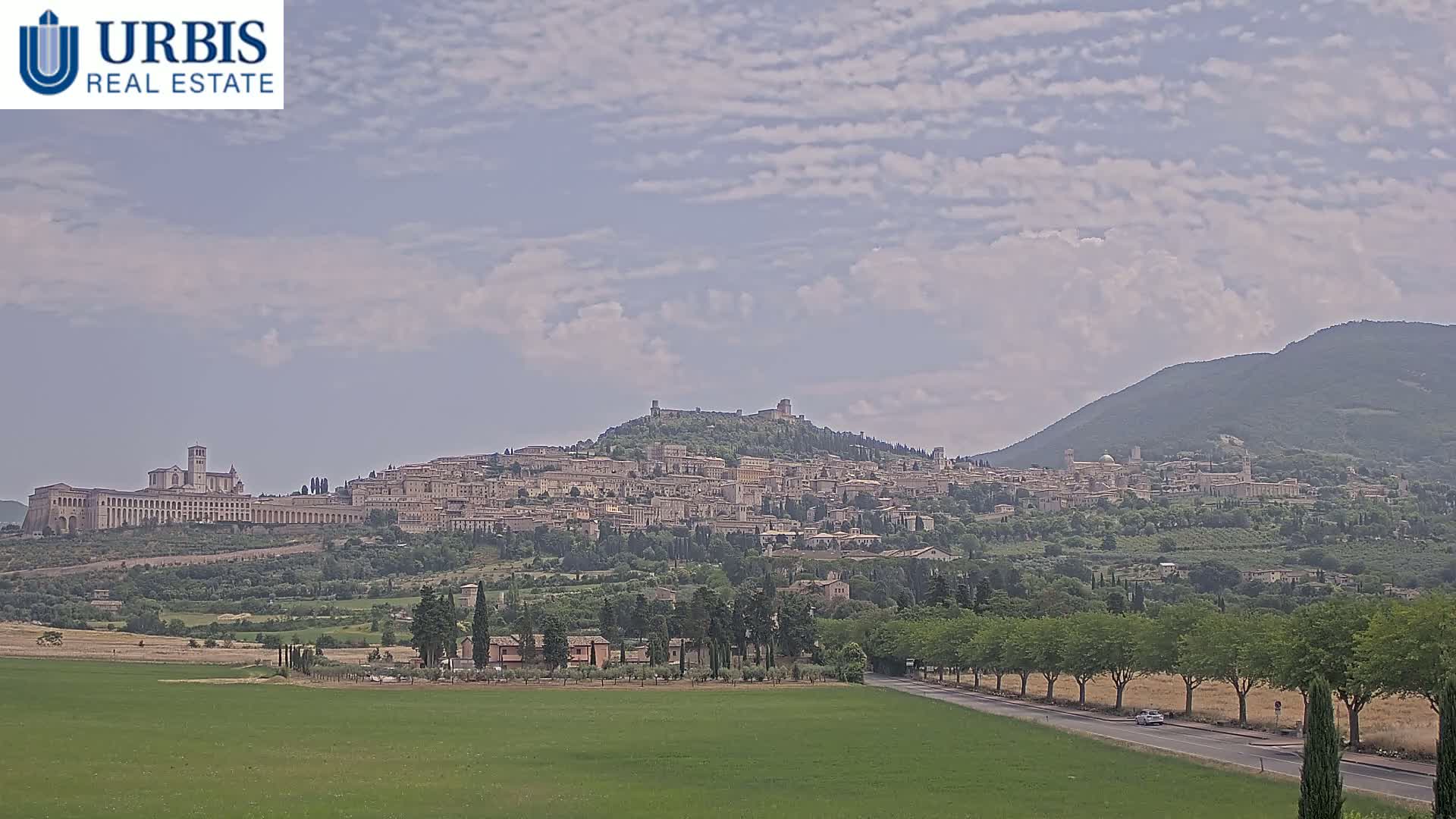 A hilltop town, nestled among green hills under a partly cloudy sky, is viewed from a distance across a flat, grassy field.