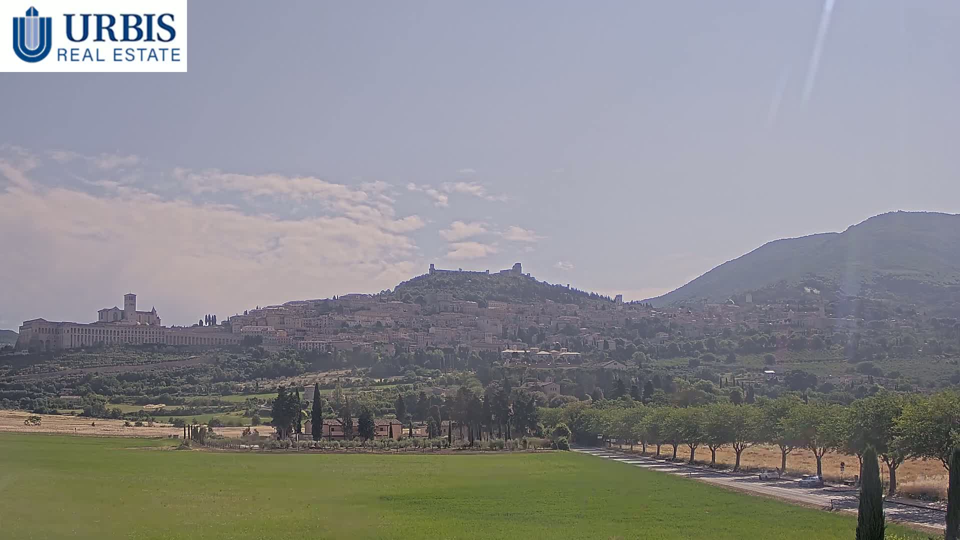 A hilltop town, including a large basilica, is visible in the distance under a partly cloudy sky, with a flat green field and tree-lined road in the foreground.