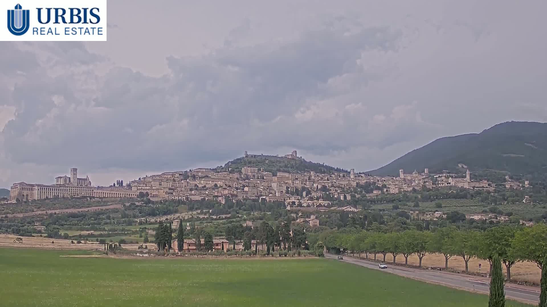 A sprawling hilltop town, under a mostly cloudy sky, is seen from a distance across a flat green field.