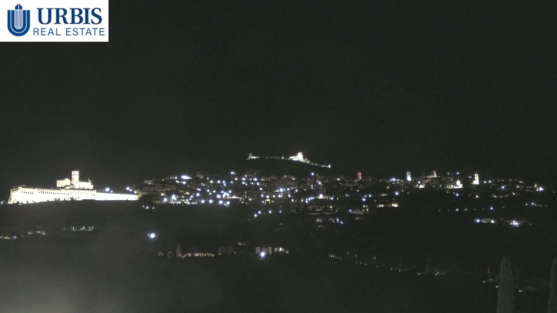 A nighttime view of a town illuminated by scattered lights, situated on a hill with a large, brightly lit structure at its base.