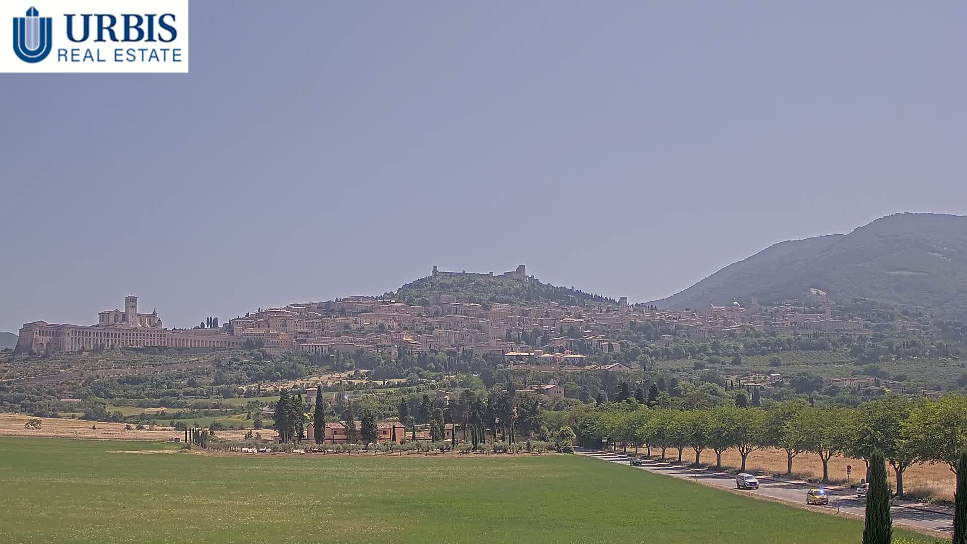 A hilltop town, nestled in a verdant valley under a clear, light-blue sky, is seen from a distance across a field.