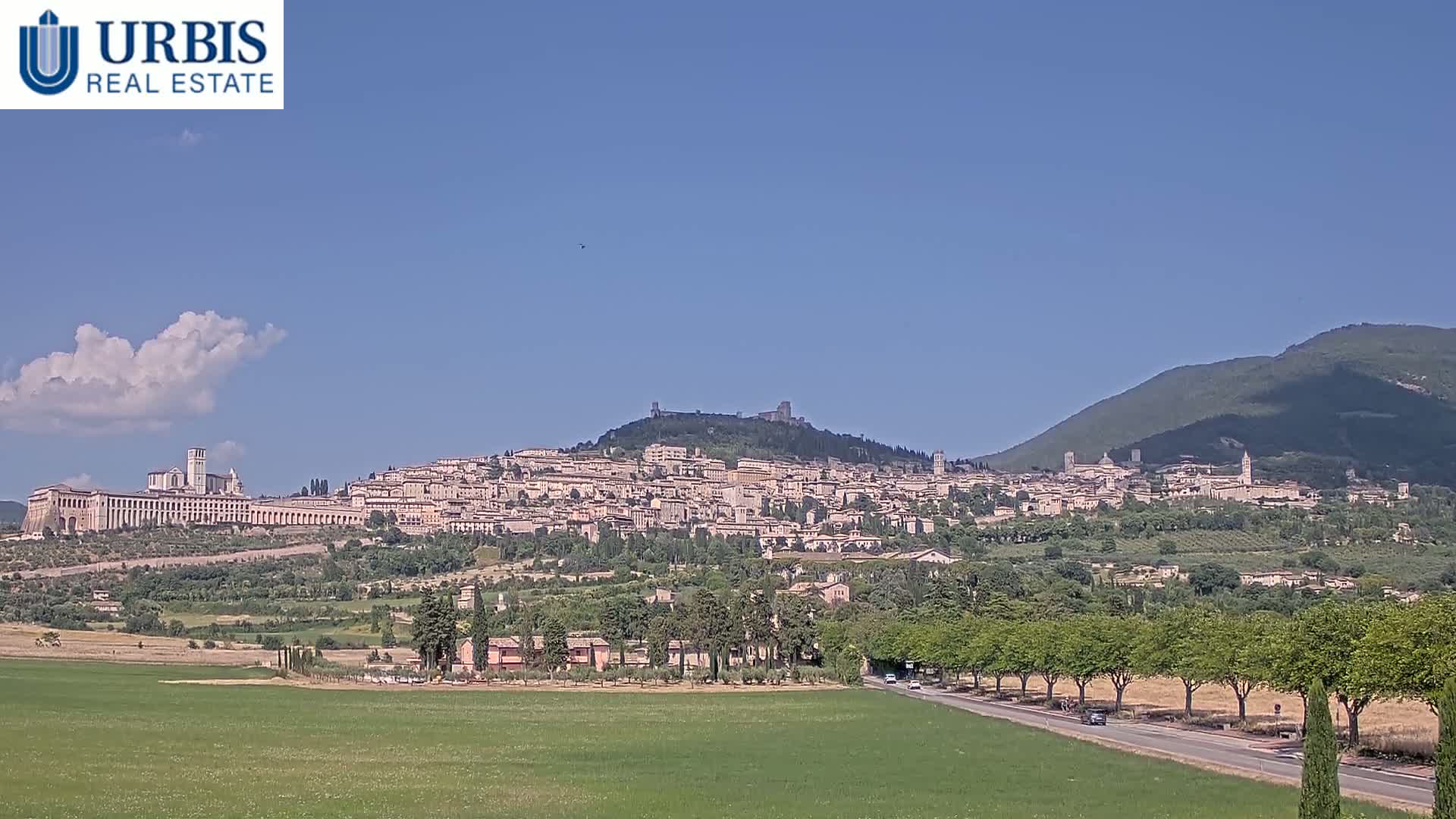 A hilltop town with many buildings, nestled among green hills under a mostly clear blue sky with a few scattered clouds.