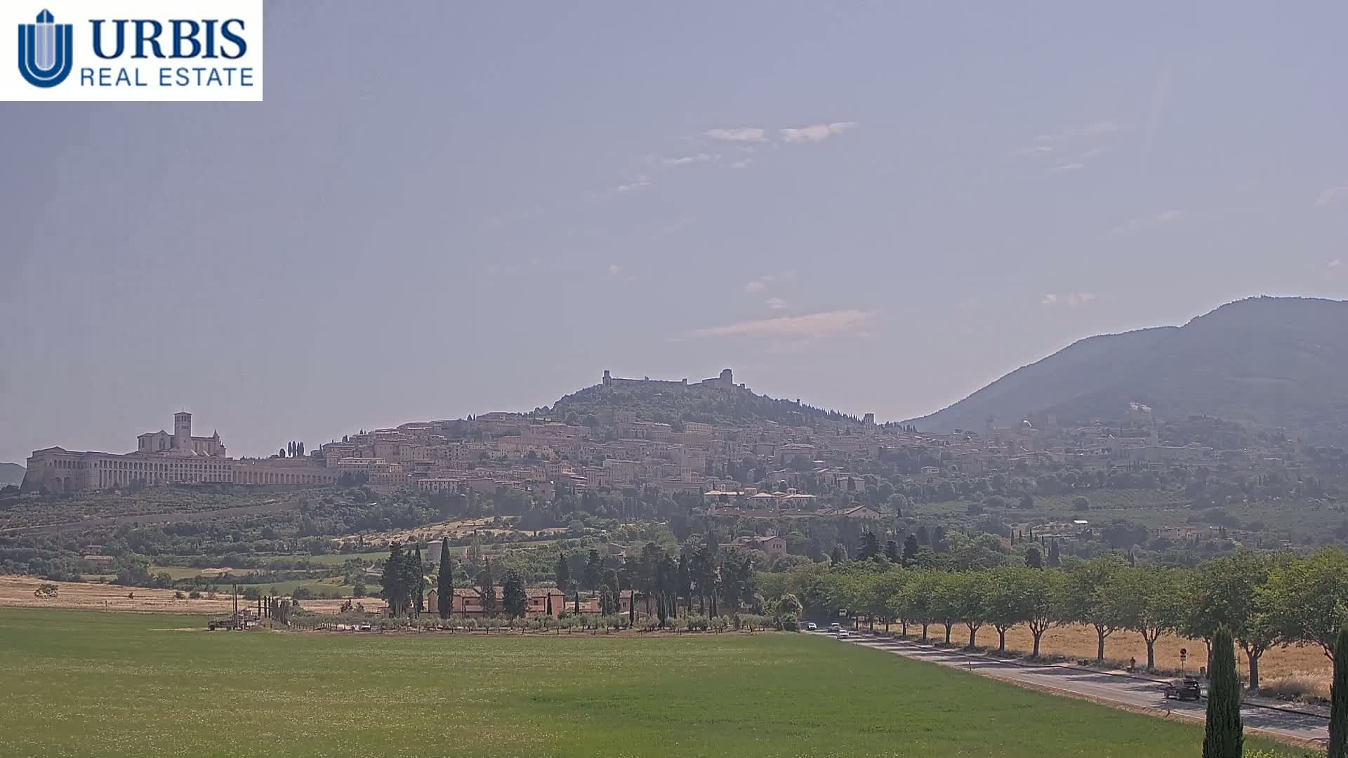 A hilltop town with a large basilica, surrounded by green fields and trees, under a hazy sky.