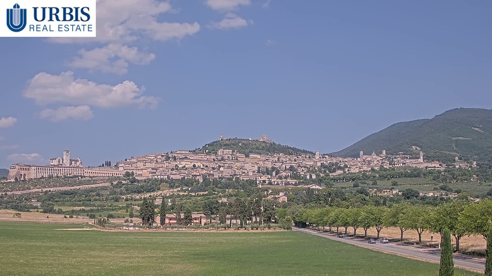 A hilltop town, nestled amongst green hills under a mostly clear blue sky, is viewed from a distance across a flat field and a tree-lined road.