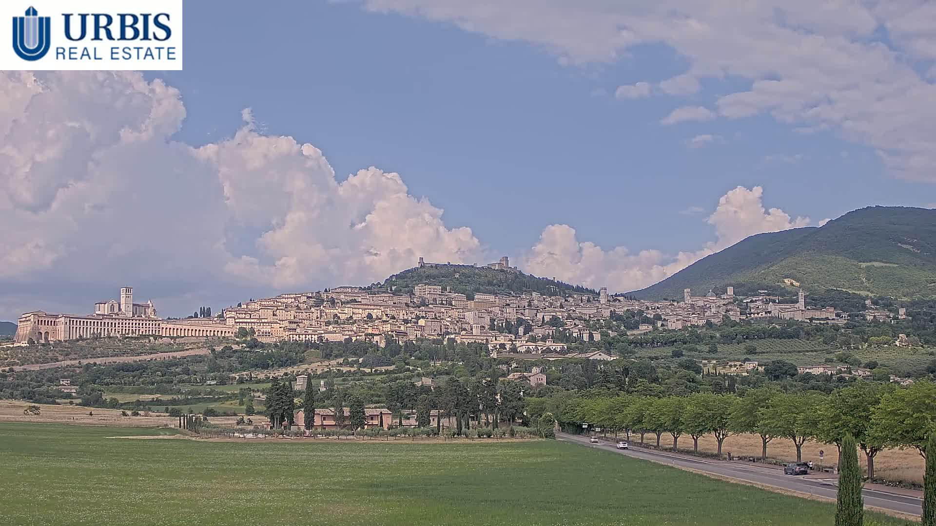 A hilltop town with a large basilica stretches across a verdant landscape under a partly cloudy sky.