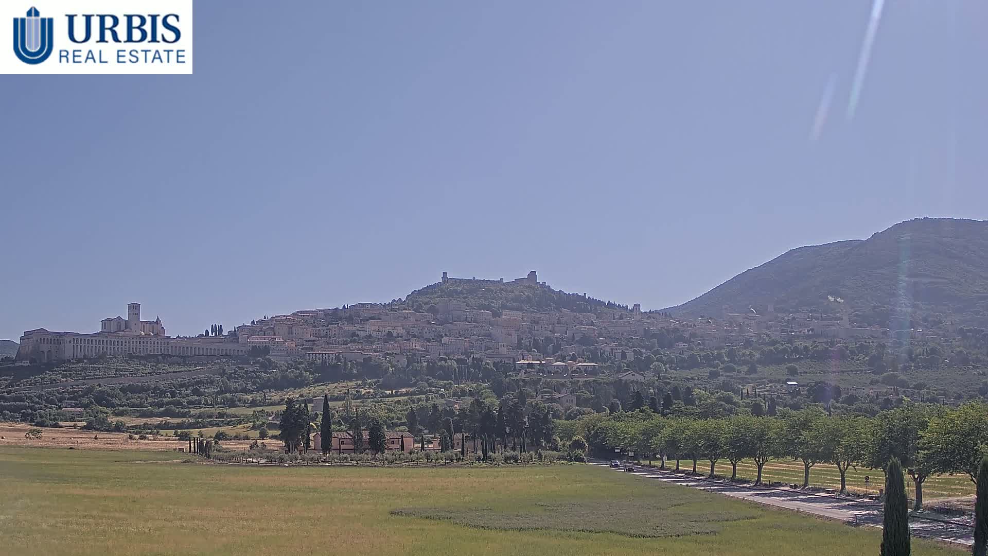 A hilltop town, possibly Assisi, is visible in the distance under a clear, sunny sky, with a large flat field in the foreground.