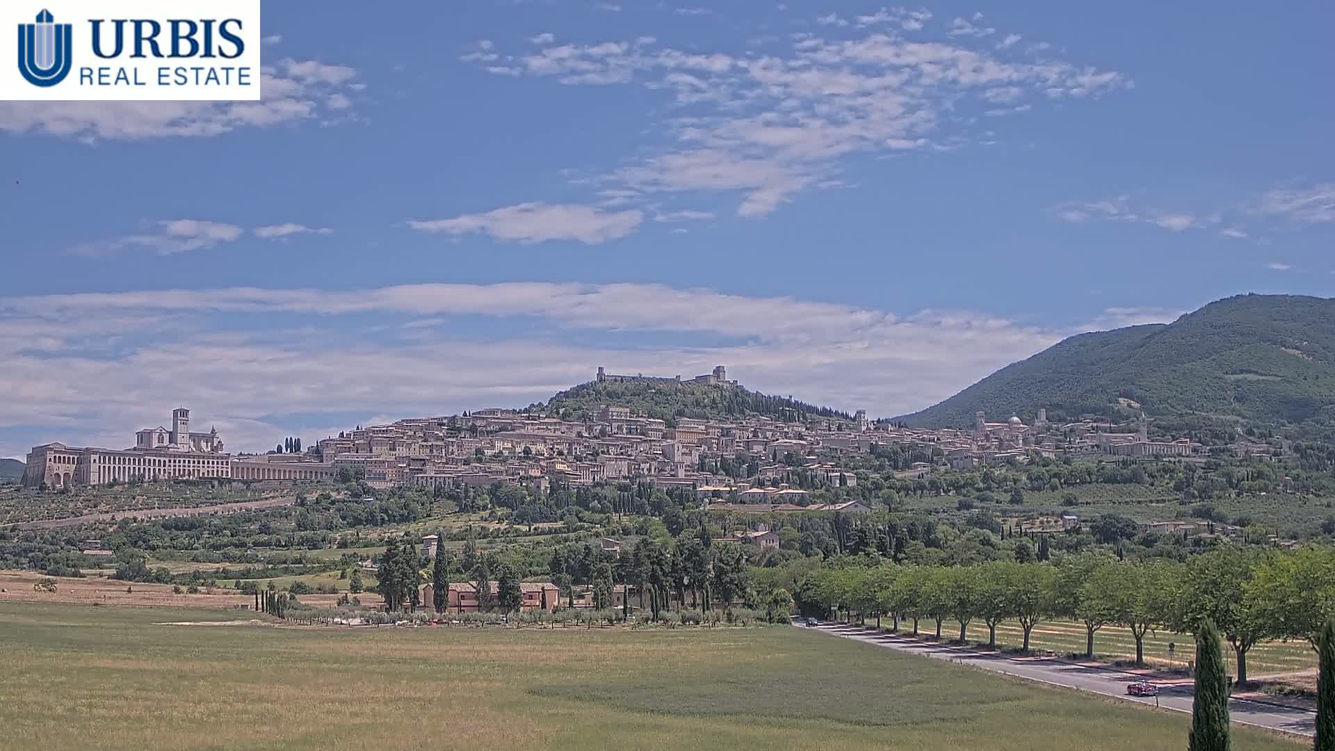 A hilltop town, possibly Assisi, is visible in the distance under a clear, sunny sky, with a large flat field in the foreground.
