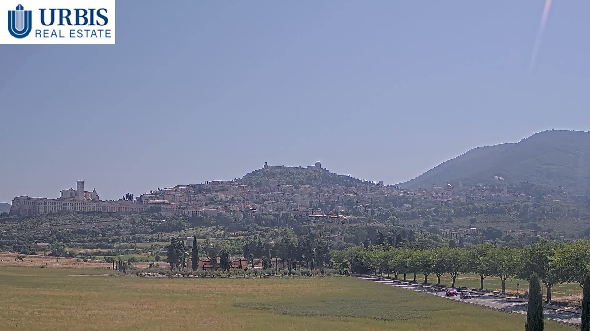 A hilltop town, partially obscured by haze, is seen from a distance on a clear, sunny day, with a verdant plain and a tree-lined road in the foreground.