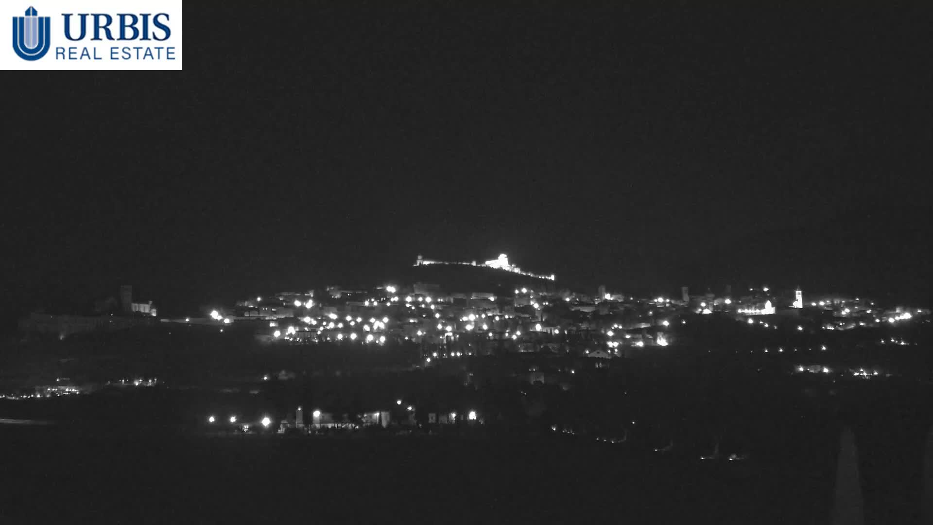 A clear night view captures a sprawling town illuminated by numerous streetlights and buildings, with a prominent, brightly lit structure atop a distant hill.