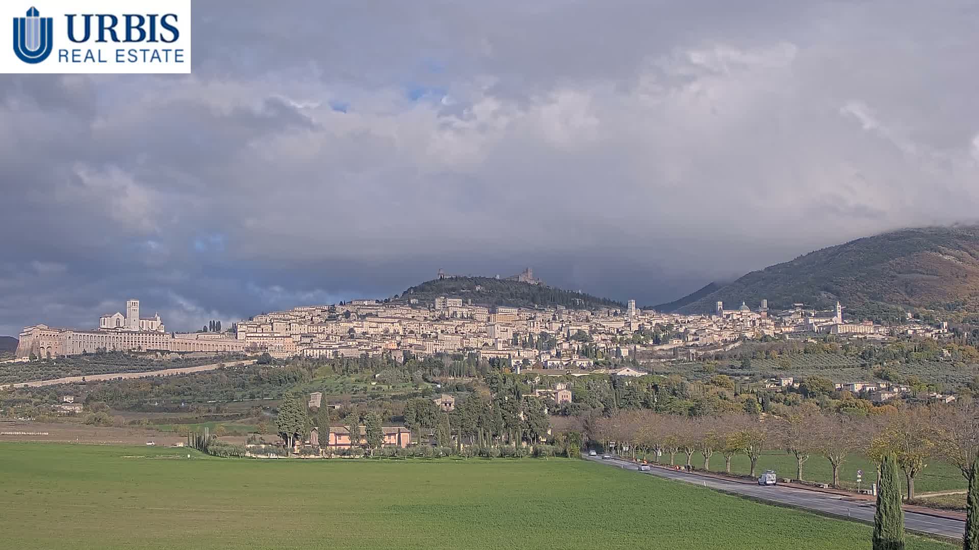 A historic Italian hillside town, likely Assisi, is visible under an overcast and misty sky, with its buildings cascading down a hill above green fields and a tree-lined road.