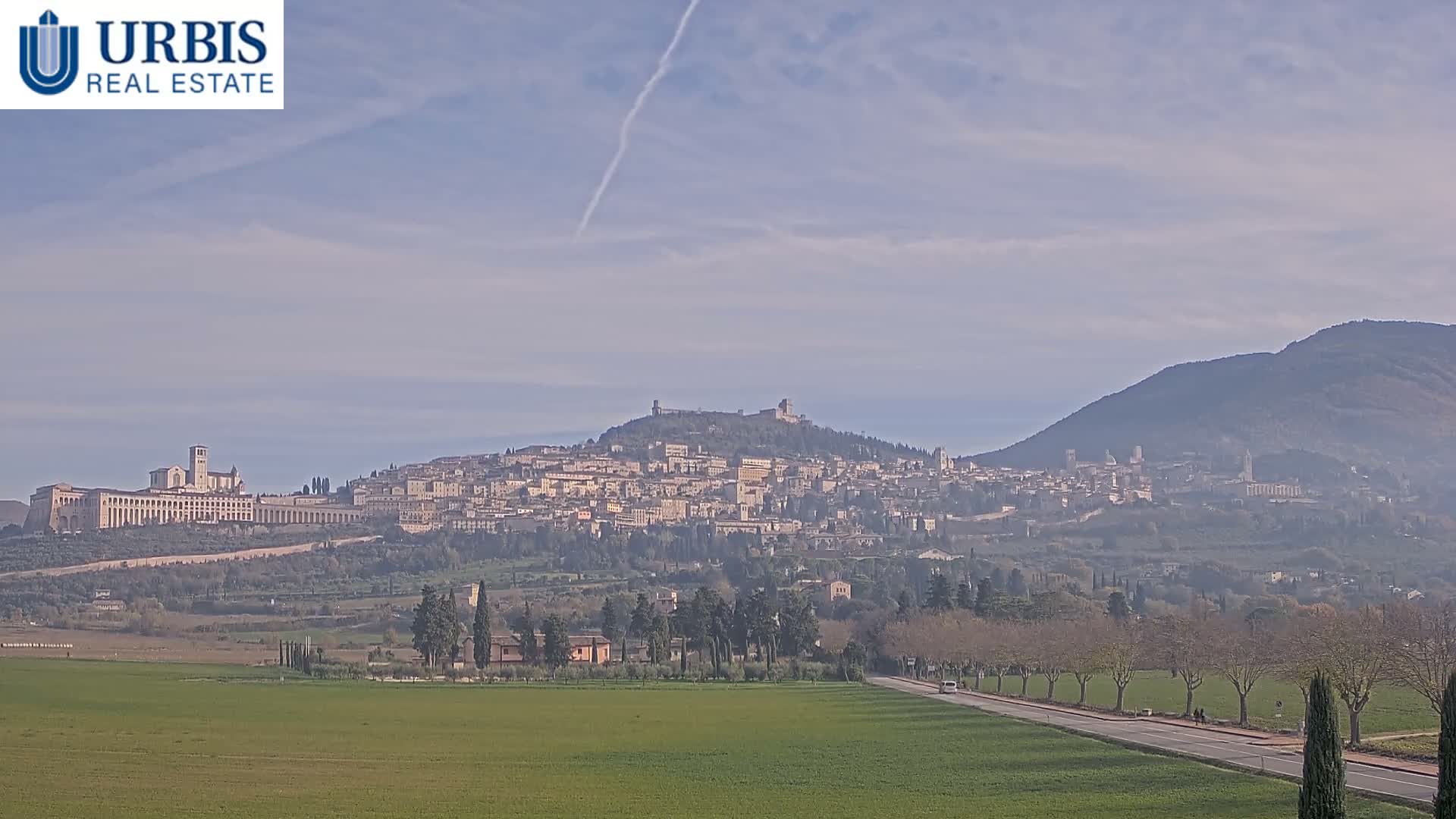 Under a clear sky with scattered clouds and a visible contrail, a historic hilltop city featuring a large basilica and a fortress overlooks green fields and a tree-lined road in the foreground.