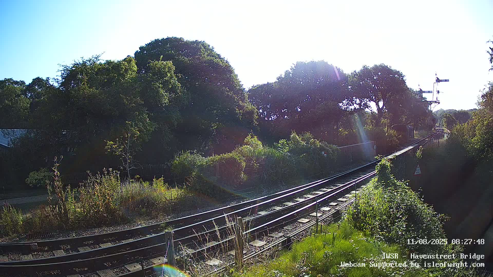 Under a bright, sunny sky, railroad tracks curve gently through a landscape of lush green vegetation and trees bordering a low brick wall.