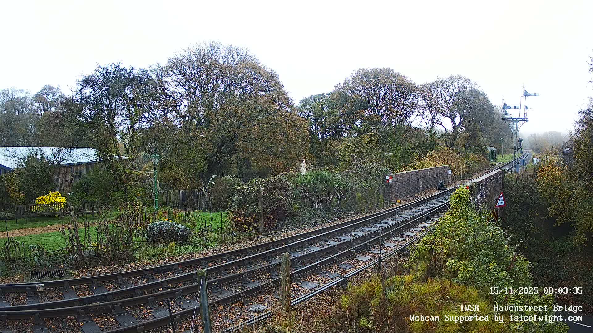Train tracks lead over a brick bridge and past a signal gantry through an autumnal landscape filled with trees, shrubs, and a garden, all under an overcast and hazy sky.