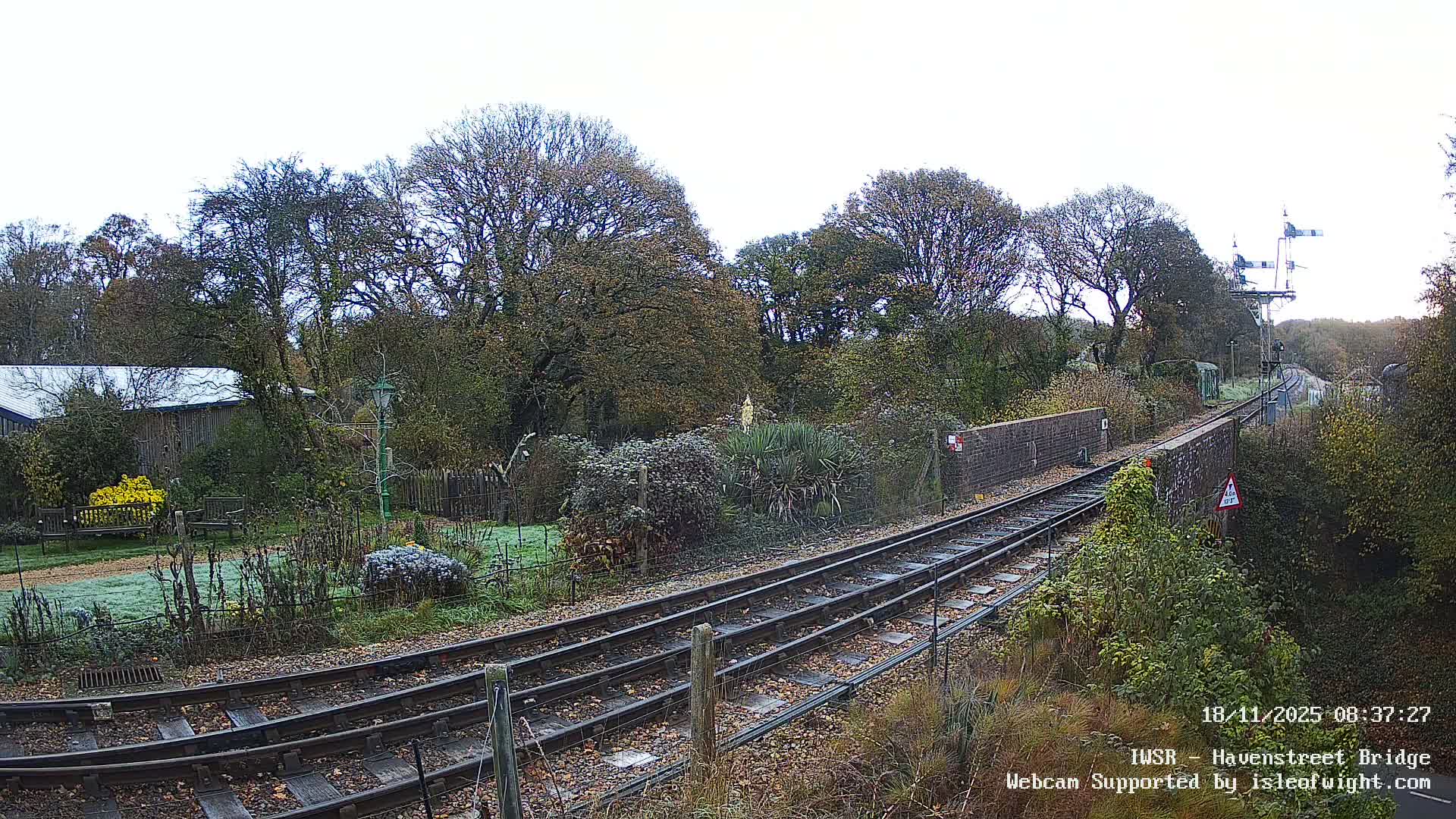 Two sets of railway tracks cut through a frost-dusted, autumnal landscape featuring a garden with bright yellow bushes, bare trees, and traditional railway signals, all under a bright, overcast sky.
