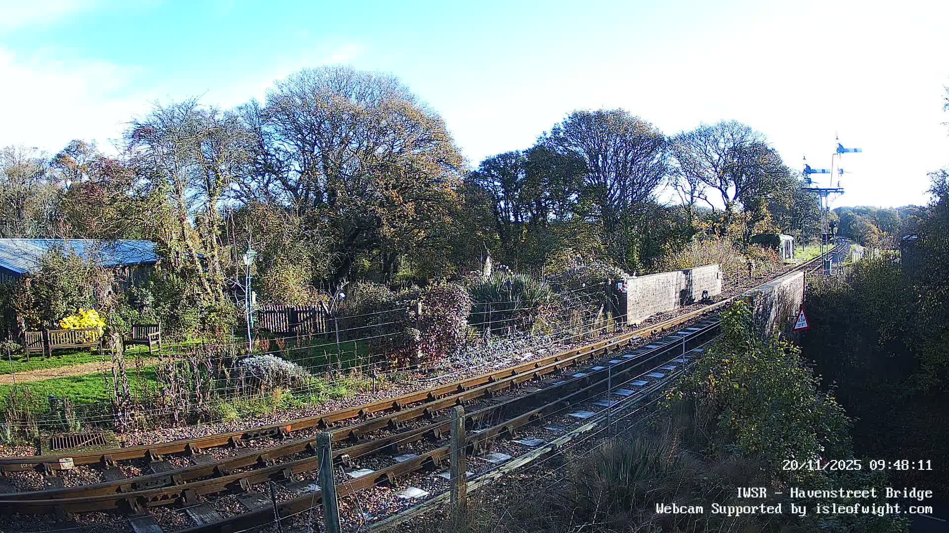 A scenic view captures railway tracks curving through a vibrant, tree-lined landscape with a garden and building to the left, all under a bright, clear blue sky on a sunny day.
