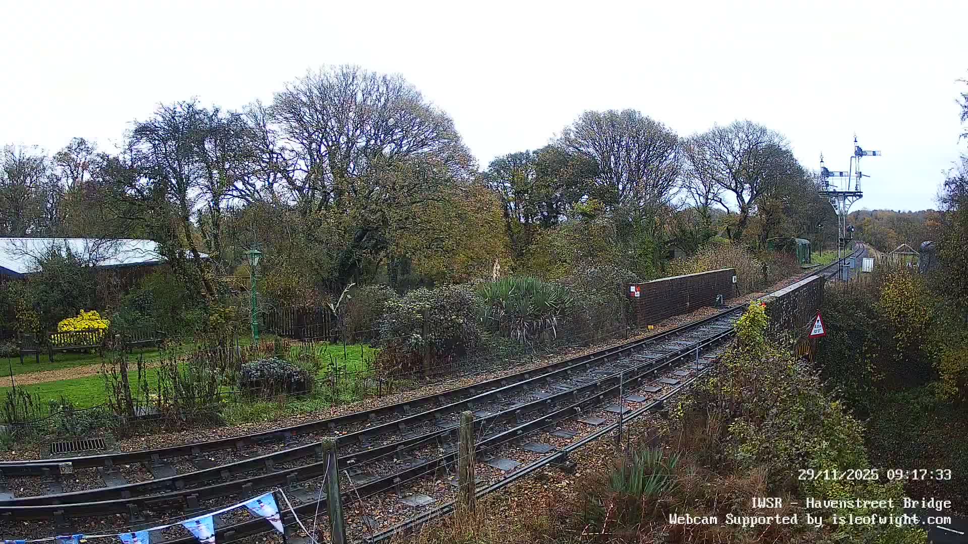 A railway line curves through a verdant, somewhat overgrown landscape featuring numerous trees, a brick wall, and a signal gantry under an overcast sky.