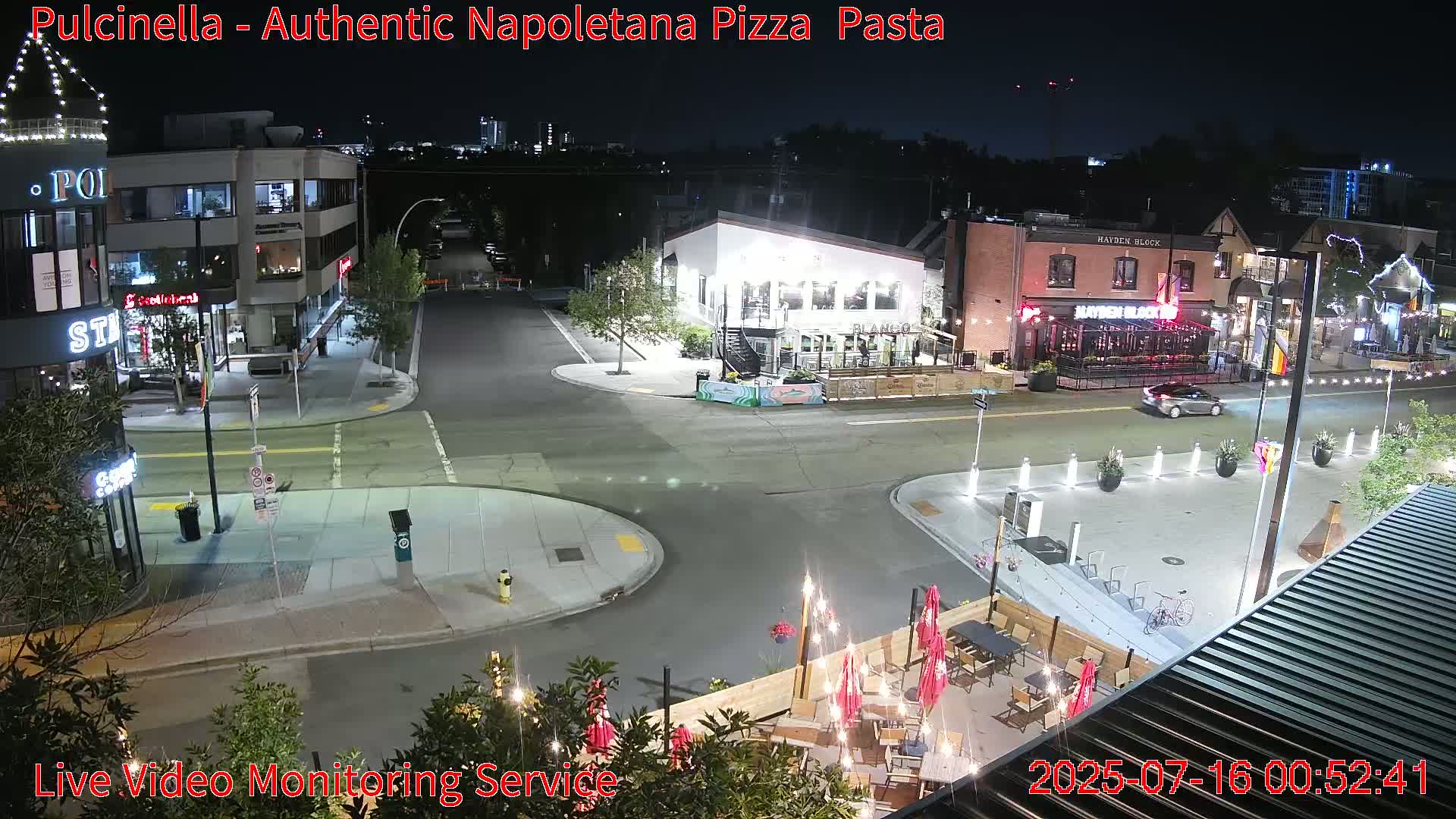 A nighttime street scene shows a mostly empty intersection with several restaurants and buildings illuminated, and an outdoor patio area with tables and string lights.