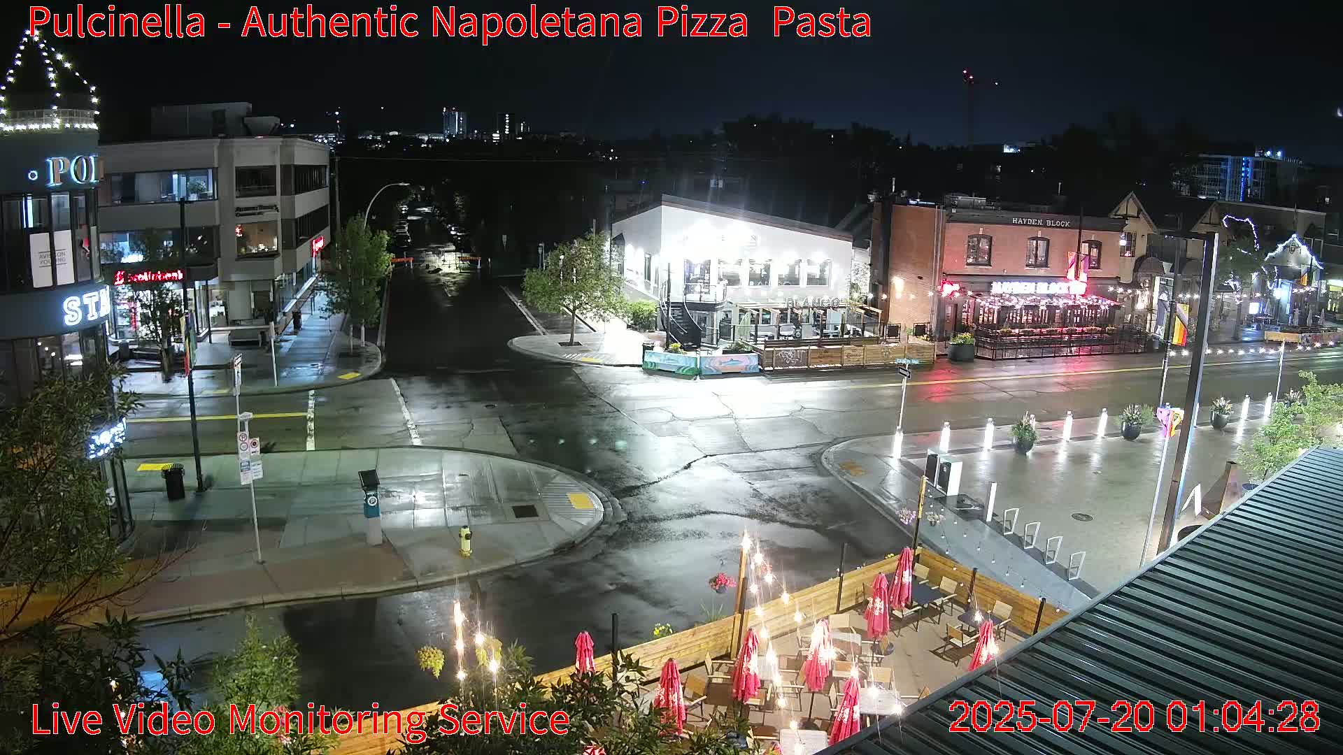 A nighttime street scene shows wet roads and several restaurants with outdoor seating under string lights, during a rainy evening.