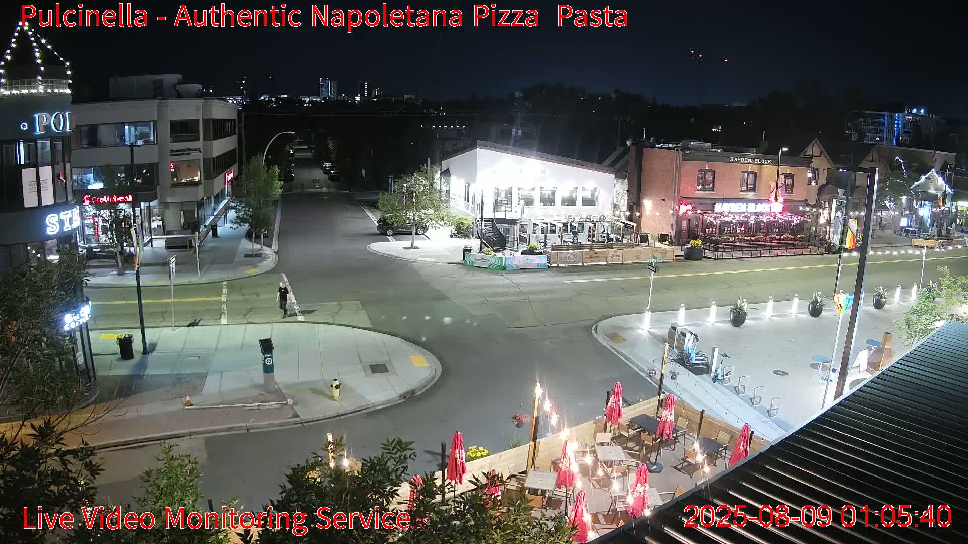 A nighttime street scene shows a pedestrian walking past restaurants with outdoor seating under illuminated umbrellas and string lights.
