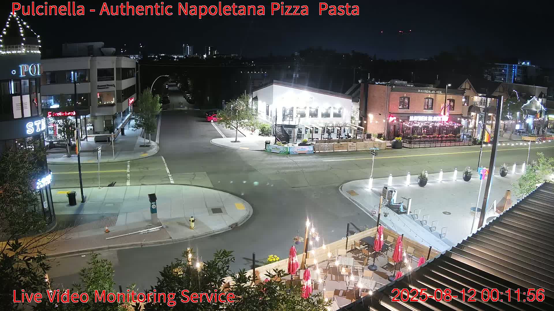 A nighttime street scene shows several illuminated buildings and a restaurant patio with tables and umbrellas under clear skies.