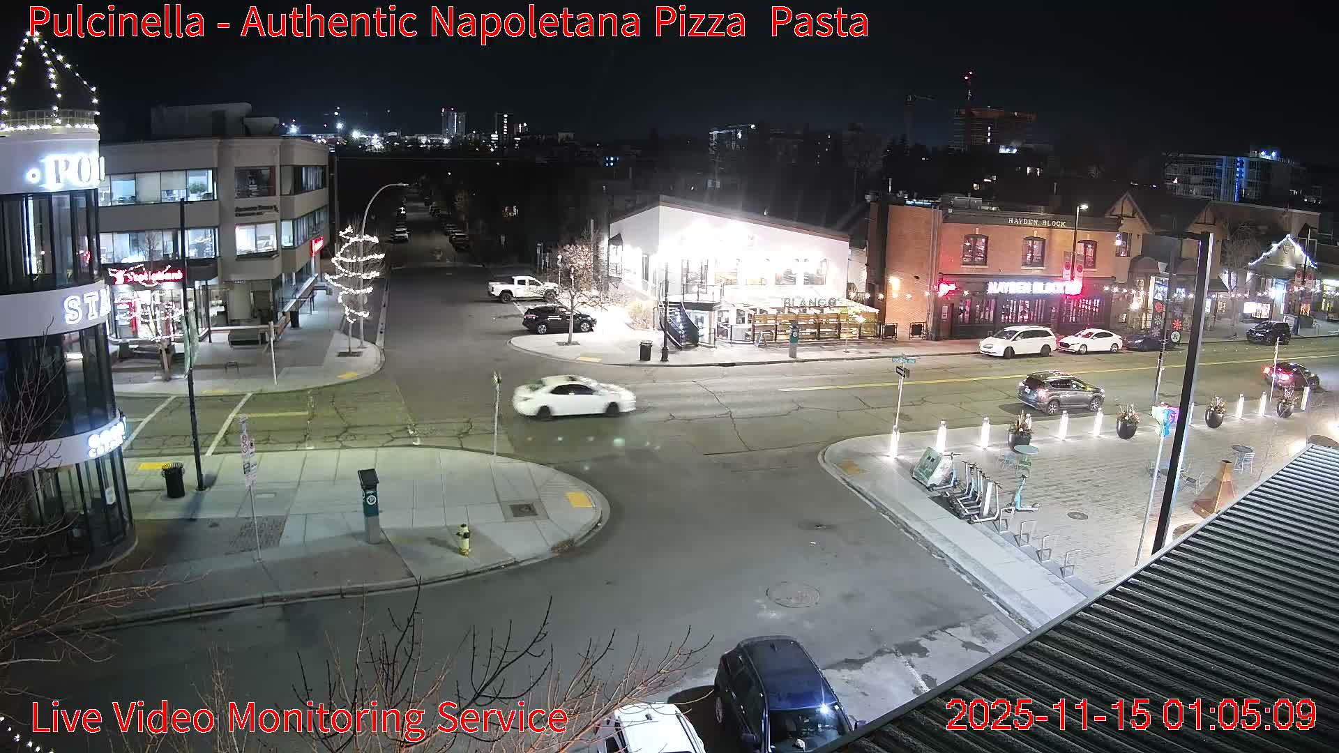 An elevated perspective captures a well-lit city intersection at night, featuring several buildings with illuminated windows, multiple cars on the streets, and decorative lights, all under a clear, dry sky.