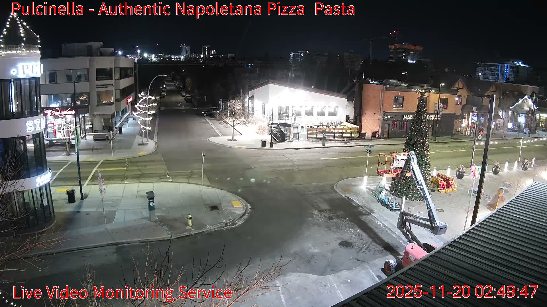 A nighttime overhead view of a city street reveals illuminated buildings and holiday decorations, including a large Christmas tree next to a boom lift, on a cool, damp evening.