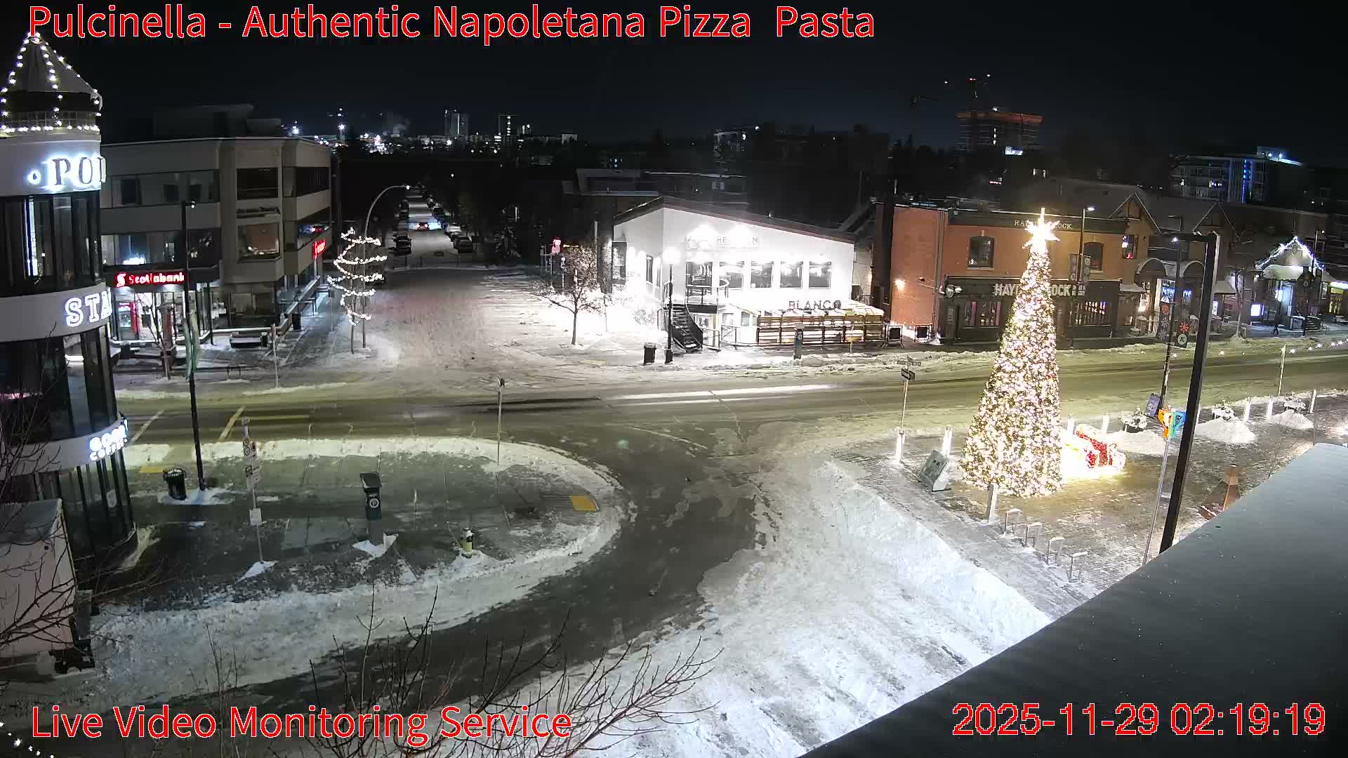 A snow-covered urban street at night is brightly illuminated by streetlights and festive decorations, prominently featuring a large, sparkling Christmas tree on the right side.