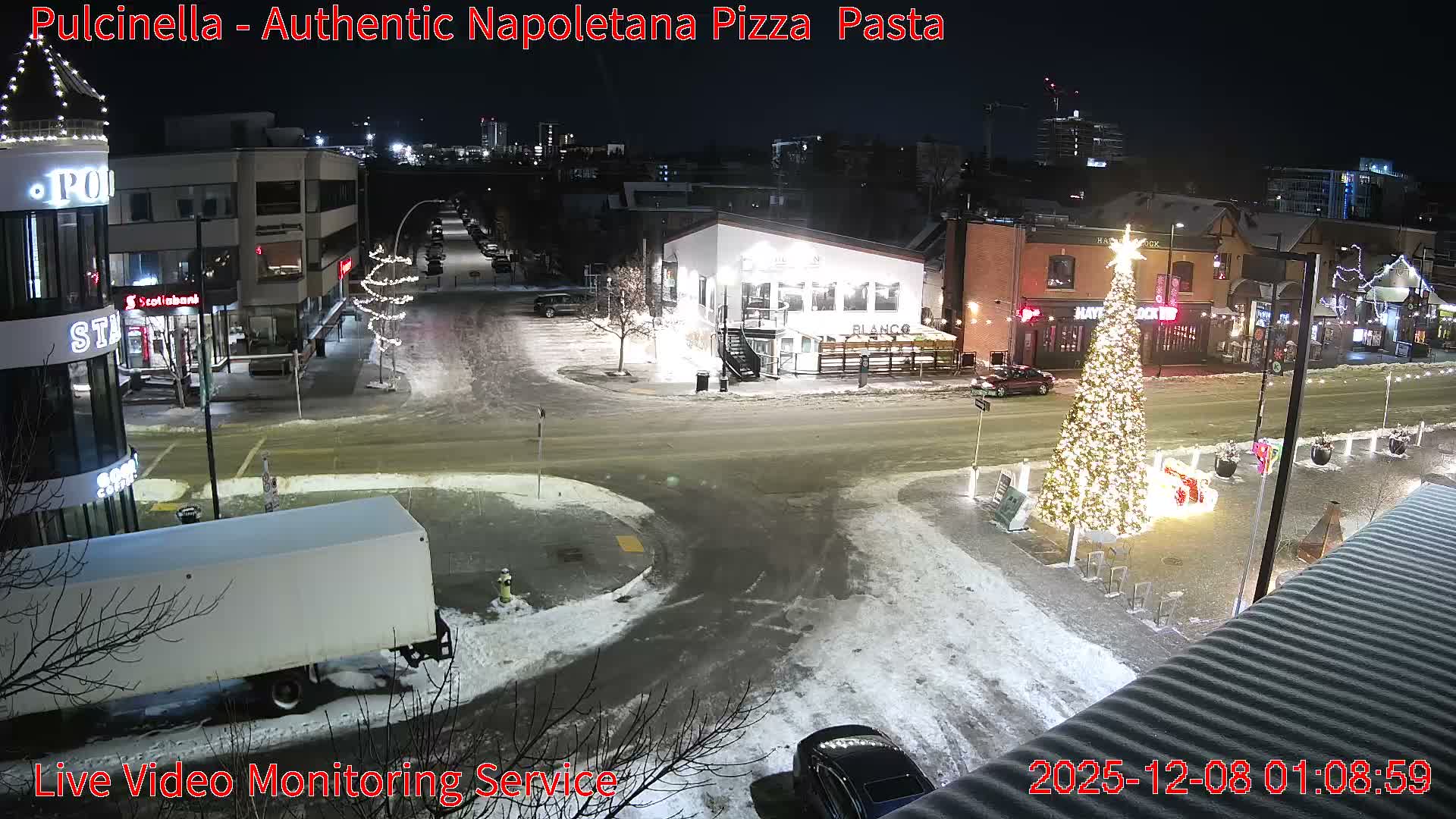 A snow-covered urban street scene at night is brightly illuminated by streetlights and extensive Christmas decorations, including a large decorated tree, with buildings and parked vehicles visible under a clear, dark sky.