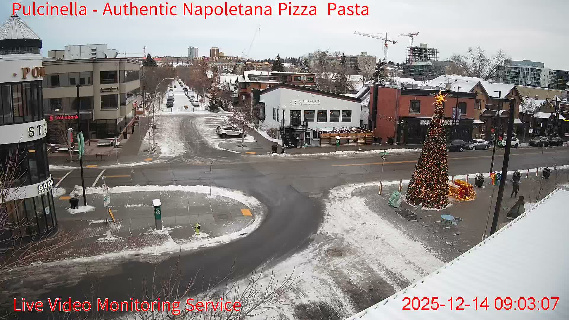 A snow-covered urban street scene at night is brightly illuminated by streetlights and extensive Christmas decorations, including a large decorated tree, with buildings and parked vehicles visible under a clear, dark sky.