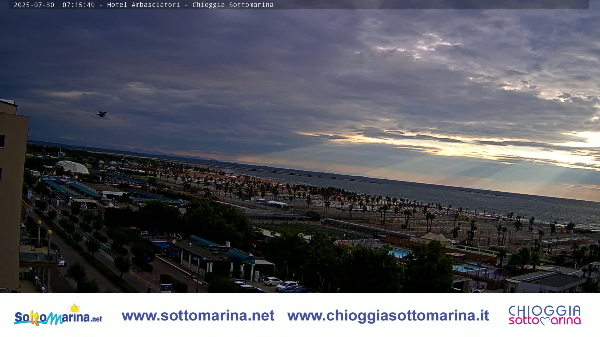 A partly cloudy day overlooks a beach with numerous umbrellas and parking lots, next to buildings and a roadway.