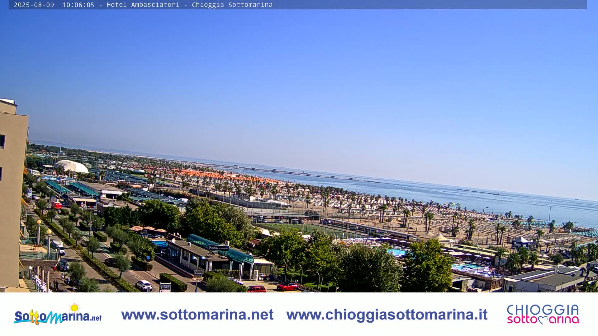 A sunny day overlooking a beach crowded with umbrellas and beachgoers, a roadway, and buildings, with calm blue water extending to the horizon.