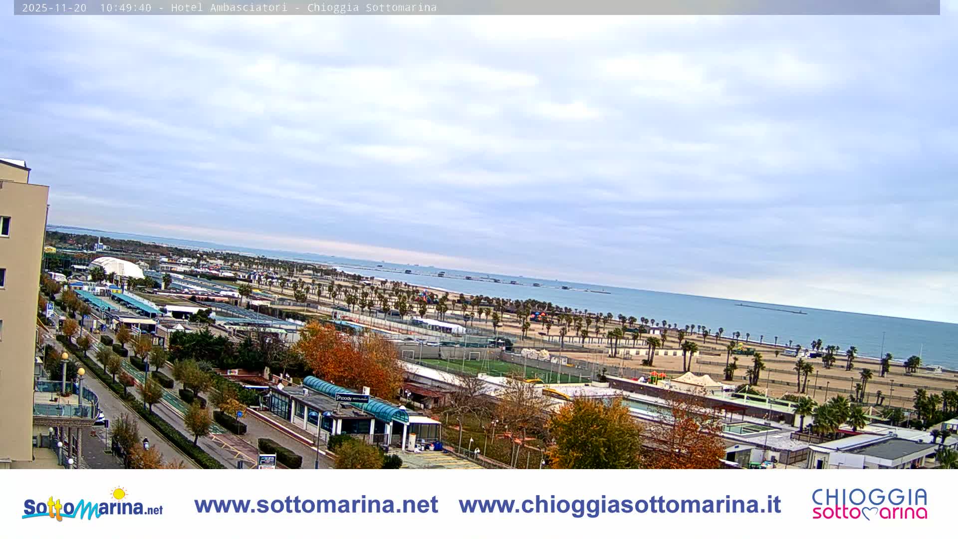 The image shows a wide, overcast view of a coastal resort town, featuring a long sandy beach lined with palm trees, various buildings, and a road with trees displaying autumn colors, all under a cloudy sky.