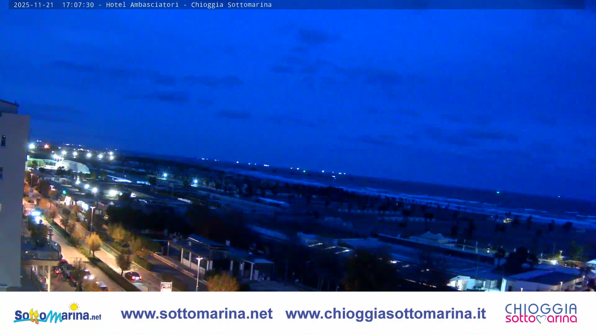 The image depicts an illuminated coastal town at dusk or night under a clear to partly cloudy deep blue sky, with streets, buildings, and the shoreline lit by numerous artificial lights.