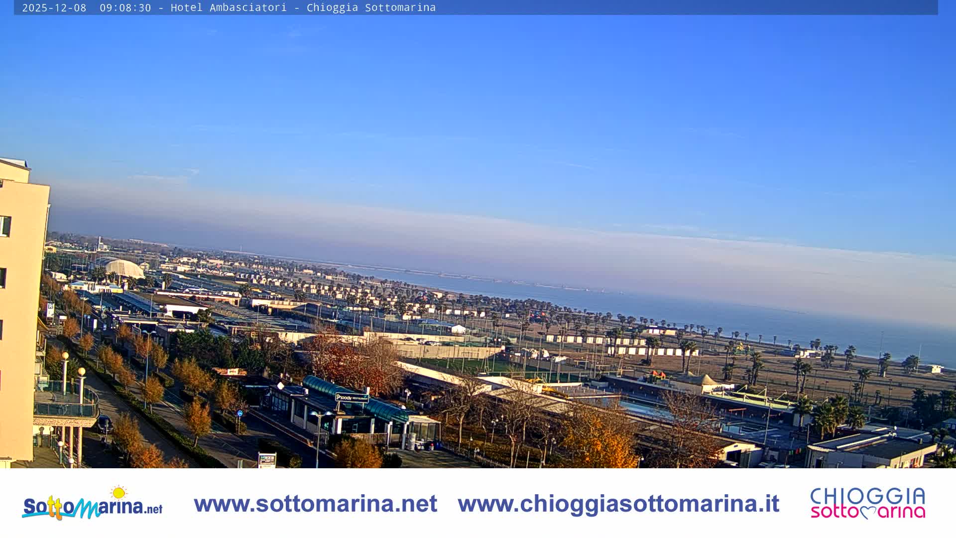 An aerial view reveals a sunny coastal town with a wide sandy beach, numerous buildings, and recreational facilities, stretching towards a calm sea under a clear blue sky.