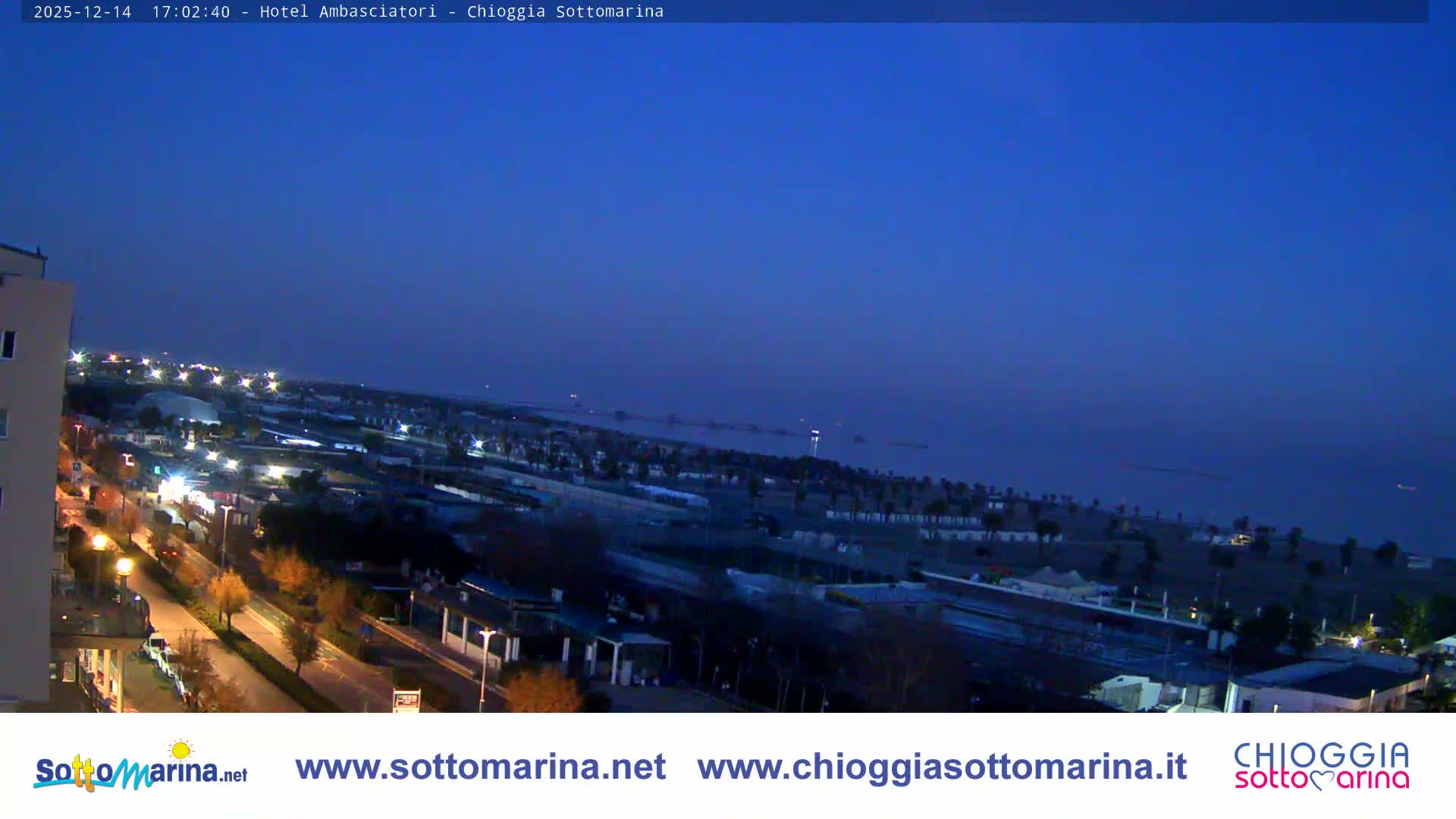 An aerial view reveals a sunny coastal town with a wide sandy beach, numerous buildings, and recreational facilities, stretching towards a calm sea under a clear blue sky.