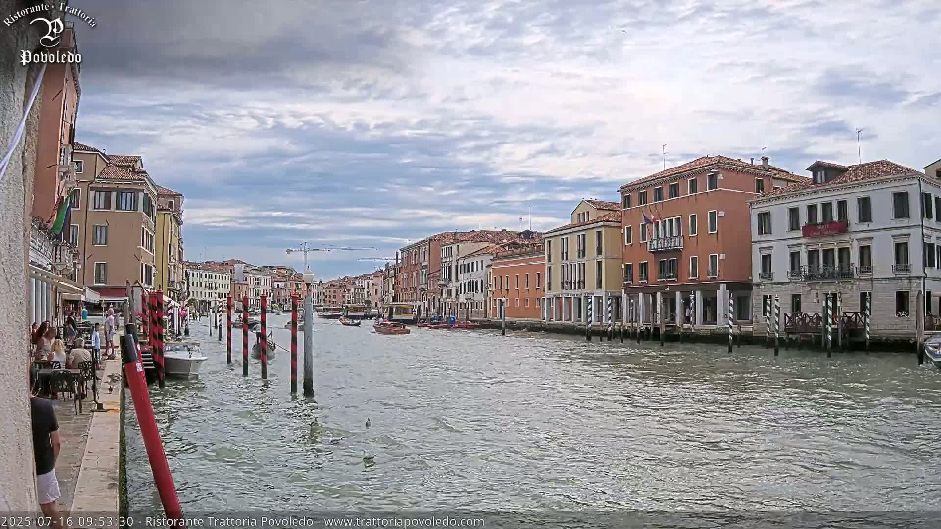 A canal in Venice, Italy, under a partly cloudy sky, shows several boats and buildings lining the waterway.